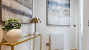 Een hal met houten vloer, een witte console tafel met een plant, een lamp en twee abstracte schilderijen aan de muur.
