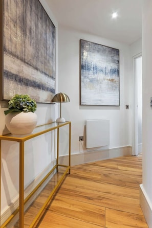 A hallway with wooden flooring, a white console table with a plant, a lamp, and two abstract paintings on the wall.