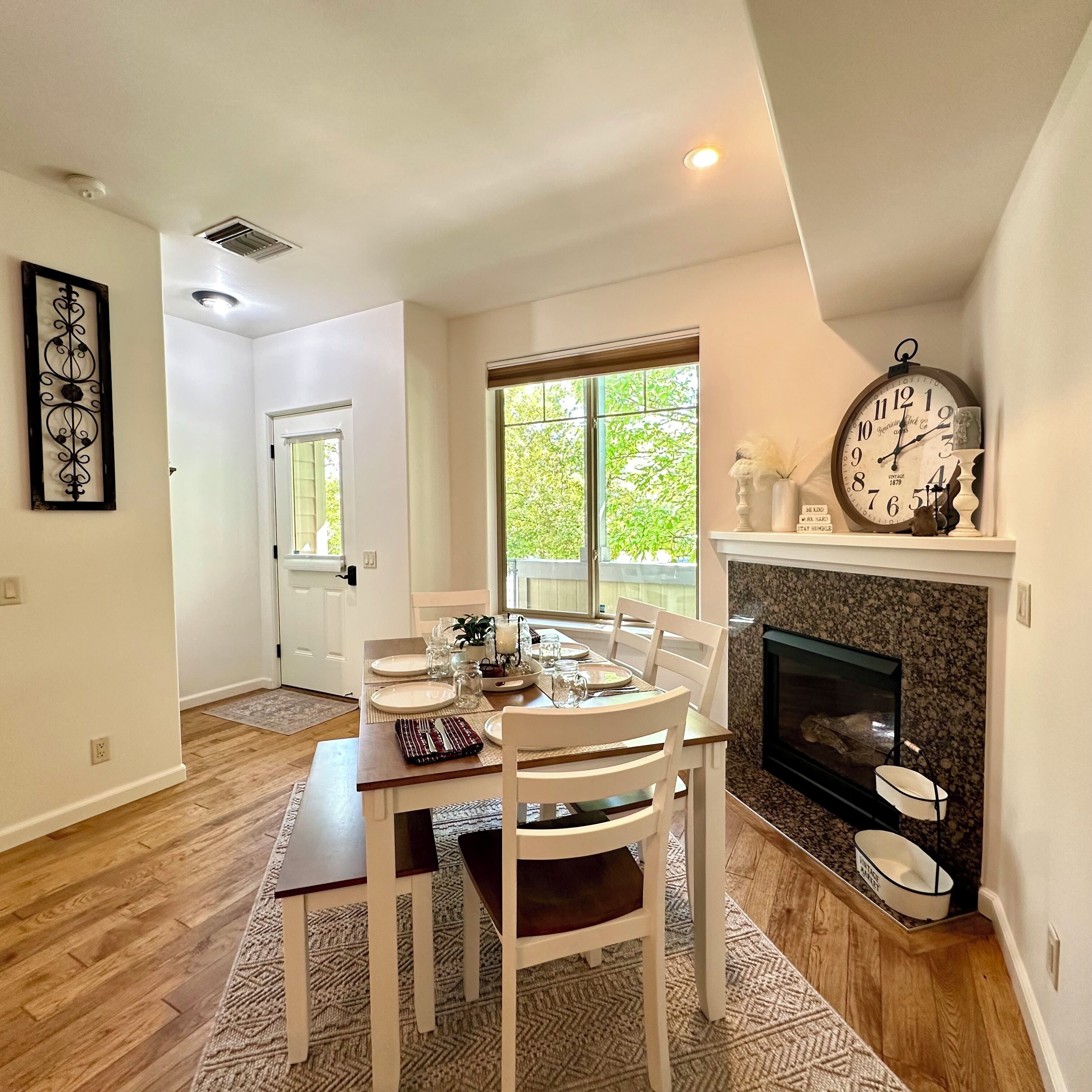 Un coin repas avec une table dressée pour quatre personnes, une cheminée et une grande horloge murale.