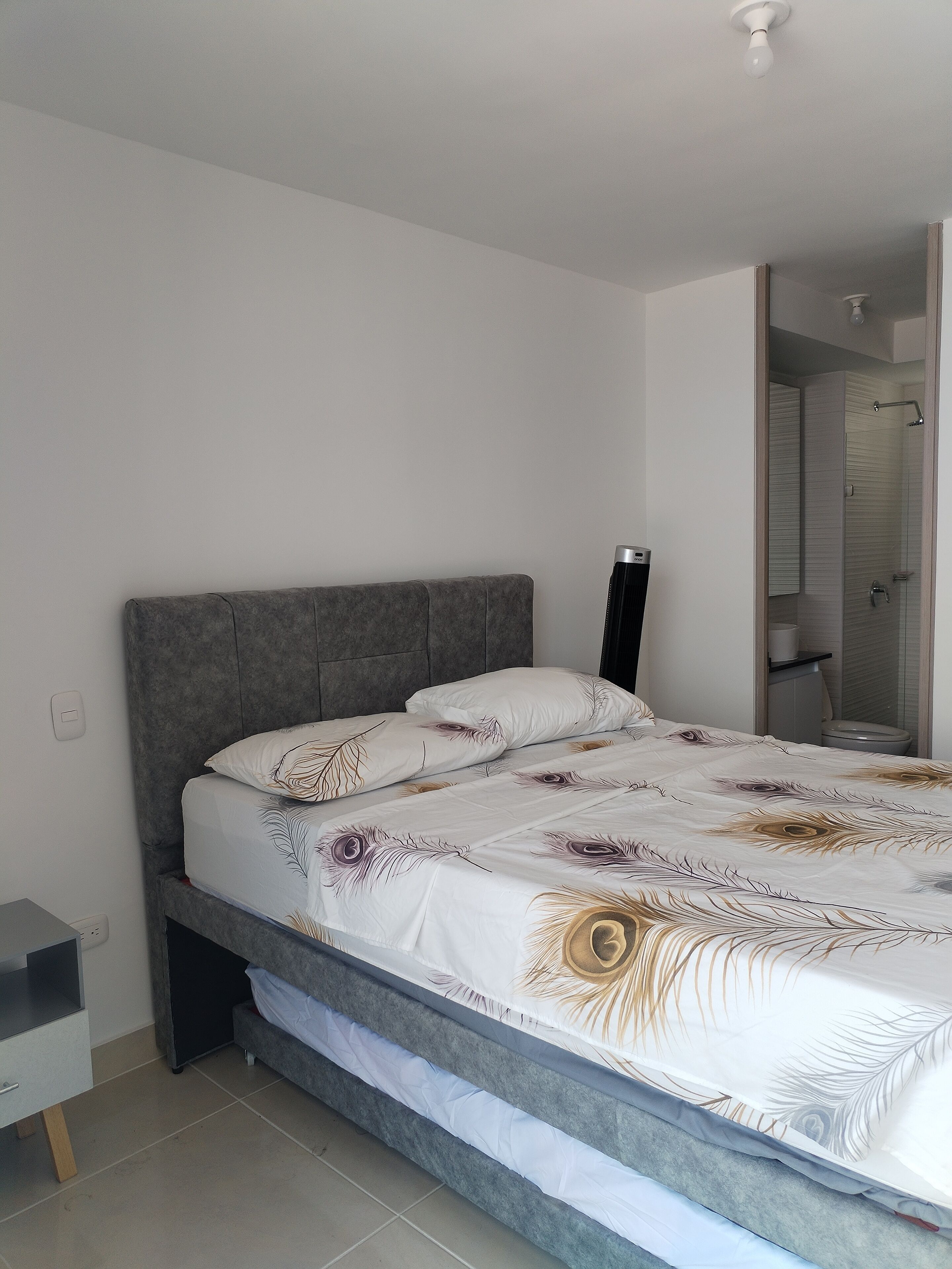 A modern bedroom with a grey upholstered headboard, a bed with white bedding featuring a feather pattern, a bedside table, and a visible bathroom door.