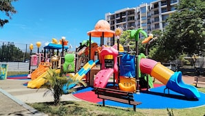 A playground with colorful slides and play structures, surrounded by a fenced area and a building in the background.