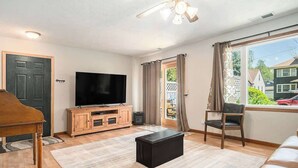 Un salon comprenant un téléviseur, un piano, un pouf noir, un meuble en bois et une grande fenêtre avec des rideaux.