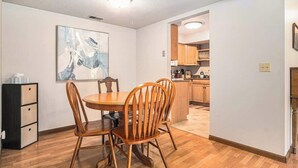 Un coin repas avec une table et des chaises en bois, un tableau accroché au mur et une cuisine en arrière-plan.
