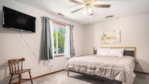 Une chambre à coucher avec un lit, un ventilateur de plafond, un téléviseur fixé au mur et une fenêtre avec des rideaux.