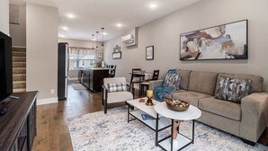 A modern living room with a sofa, coffee table, and TV stand. There is a kitchen area in the background with a dining table and chairs. - Trendy Townhouse in Midtown Omaha|Rooftop Retreat (Omaha)