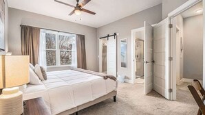 A bedroom with a bed, a ceiling fan, a window with curtains, and a door leading to another room. - Trendy Townhouse in Midtown Omaha|Rooftop Retreat (Omaha)