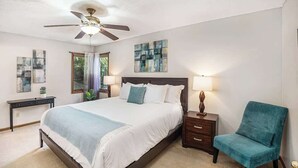 Ein Schlafzimmer mit Bett, Deckenventilator, Nachttisch mit Lampe, Stuhl und Bildern an den Wänden.