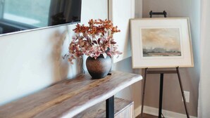 A modern living room with a wooden console table, a vase with dried flowers, a framed painting on easel, and a mounted TV.