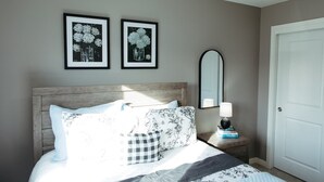 A bedroom with a wooden headboard, a bed with patterned bedding, two framed floral pictures on the wall, a bedside table with a lamp, and a door.