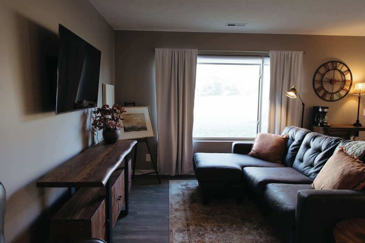 Ein Wohnzimmer mit einem dunkelbraunen Ledersofa, einem Holztisch, einem flachbildfernseher an der Wand, einem Gemälde auf einem Stativ und einem großen Fenster mit Vorhängen.