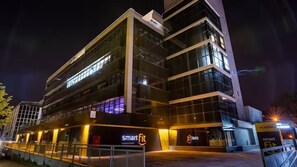 Un edificio moderno con fachada de vidrio, identificado como "Smart Fit", y un letrero que dice "Acesso".