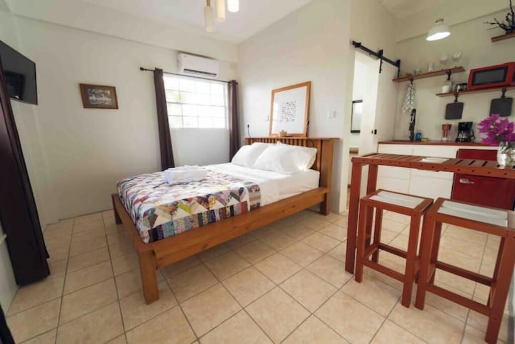 A bedroom with a wooden bed, a desk, and a chair. There is a window with curtains, a wall-mounted air conditioner, and a shelf with various items.