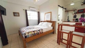 A bedroom with a wooden bed, a desk, and a chair. There is a window with curtains, a wall-mounted air conditioner, and a shelf with various items. - Irie Heights Oceanview (Gros Islet)