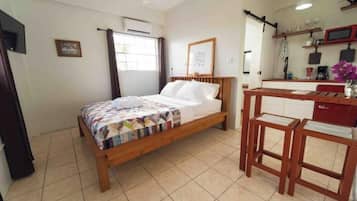 A bedroom with a wooden bed, a desk, and a chair. There is a window with curtains, a wall-mounted air conditioner, and a shelf with various items.