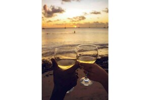 Dos manos sosteniendo copas de vino en una playa con un atardecer de fondo.