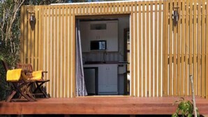 A wooden cabin with a kitchen area visible through an open door, a deck with a chair, and a small table. - Unique Container Home with Open Air Bathroom (Gros Islet)
