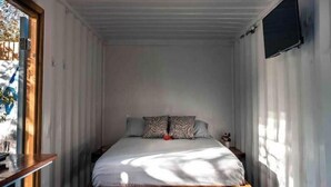 A small, single-bed room with a wooden headboard, a bedside table, and a window with curtains. - Unique Container Home with Open Air Bathroom (Gros Islet)