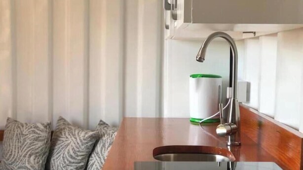 A modern kitchen with a stainless steel sink, a green and white container, and patterned pillows on a dark sofa. - Unique Container Home with Open Air Bathroom (Gros Islet)