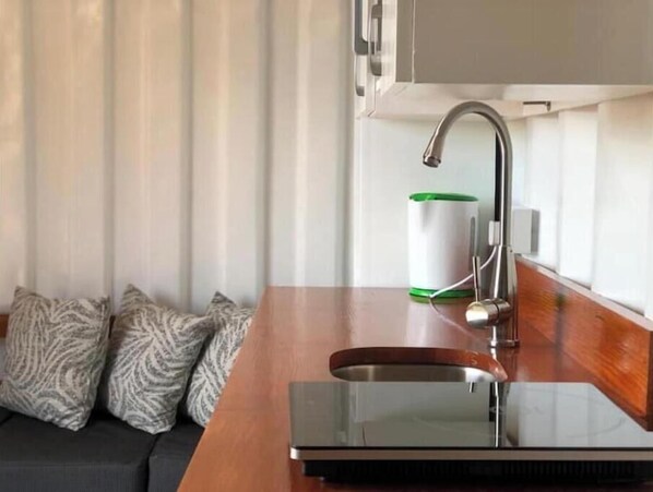 A modern kitchen with a stainless steel sink, a green and white container, and patterned pillows on a dark sofa.