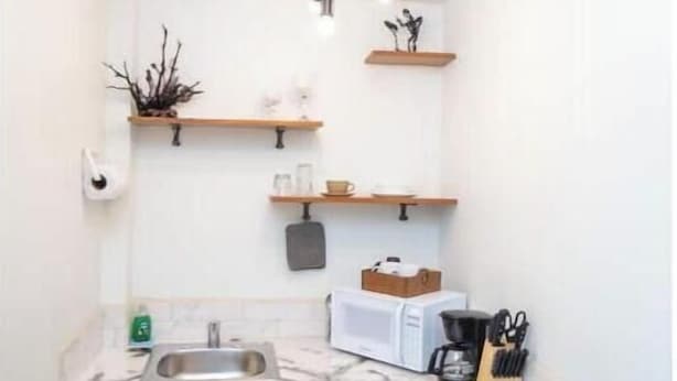 Eine kompakte Küche mit weißen Schränken, einer Spüle, einer Mikrowelle, einer Kaffeemaschine und einem Herd. Über der Arbeitsplatte befinden sich Holzregale und eine Deckenleuchte.