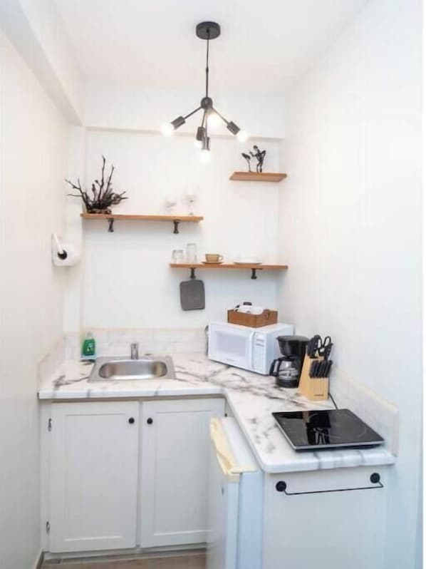 A compact kitchen with white cabinets, a sink, a microwave, a coffee maker, and a stove. There are wooden shelves above the counter and a hanging light fixture.
