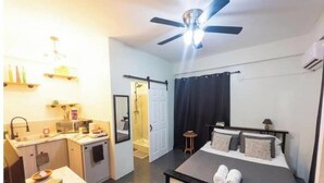 A compact hotel room with a bed, a desk, a chair, a small table, a ceiling fan, and a shower area. - Irie Heights Reggae Studio (Gros Islet)