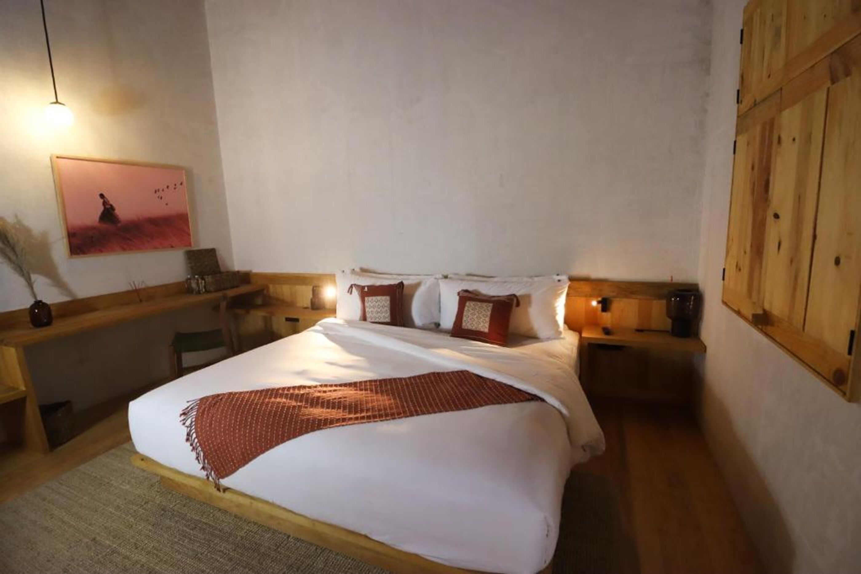 A bedroom with a bed, wooden headboard, bedside tables, a nightstand, a lamp, a framed picture on the wall, and a wooden wardrobe.