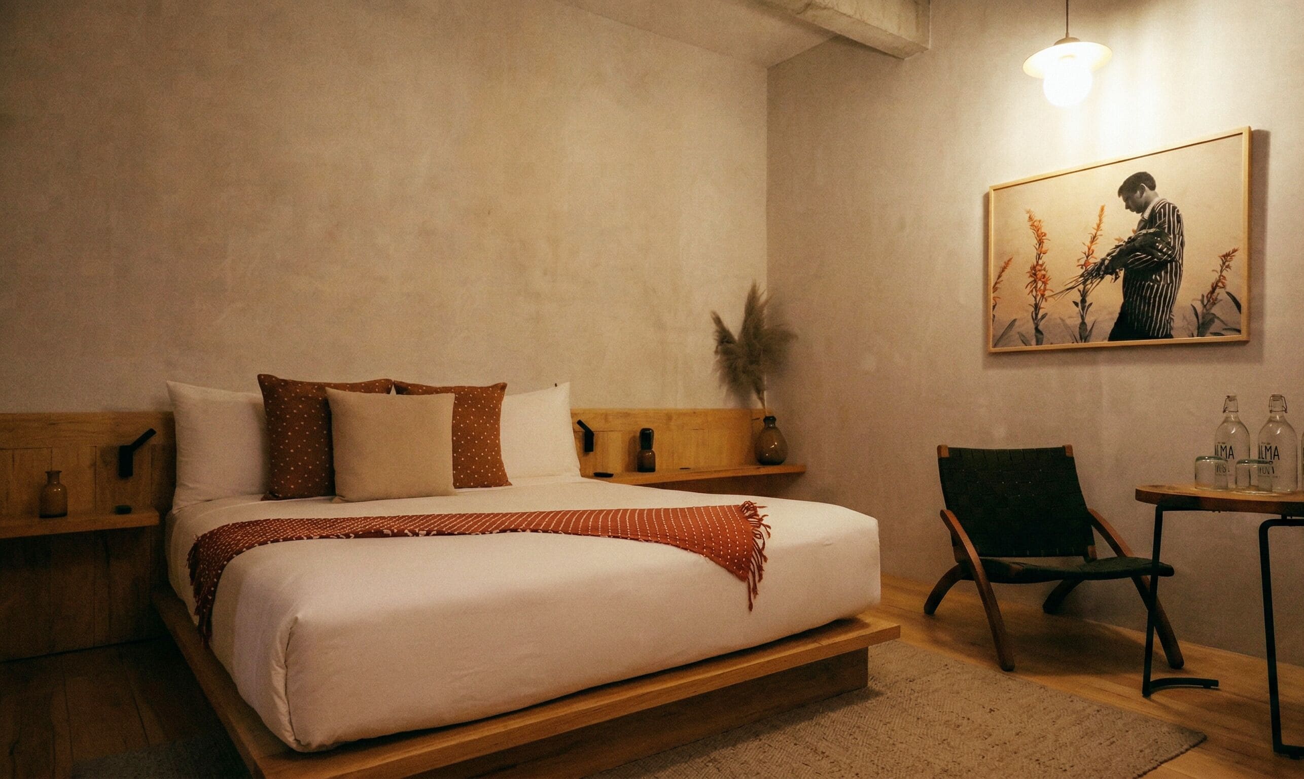 A bedroom with a bed, pillows, a chair, a small table with bottles, a wall-mounted painting, and a hanging light fixture.