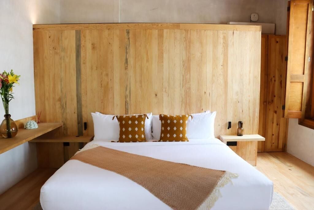A bedroom with a wooden headboard, a large bed with white bedding and patterned pillows, a bedside table with a vase of flowers, and a wooden nightstand.