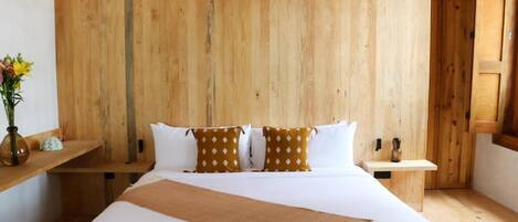 A bedroom with a wooden headboard, a large bed with white bedding and patterned pillows, a bedside table with a vase of flowers, and a wooden nightstand.