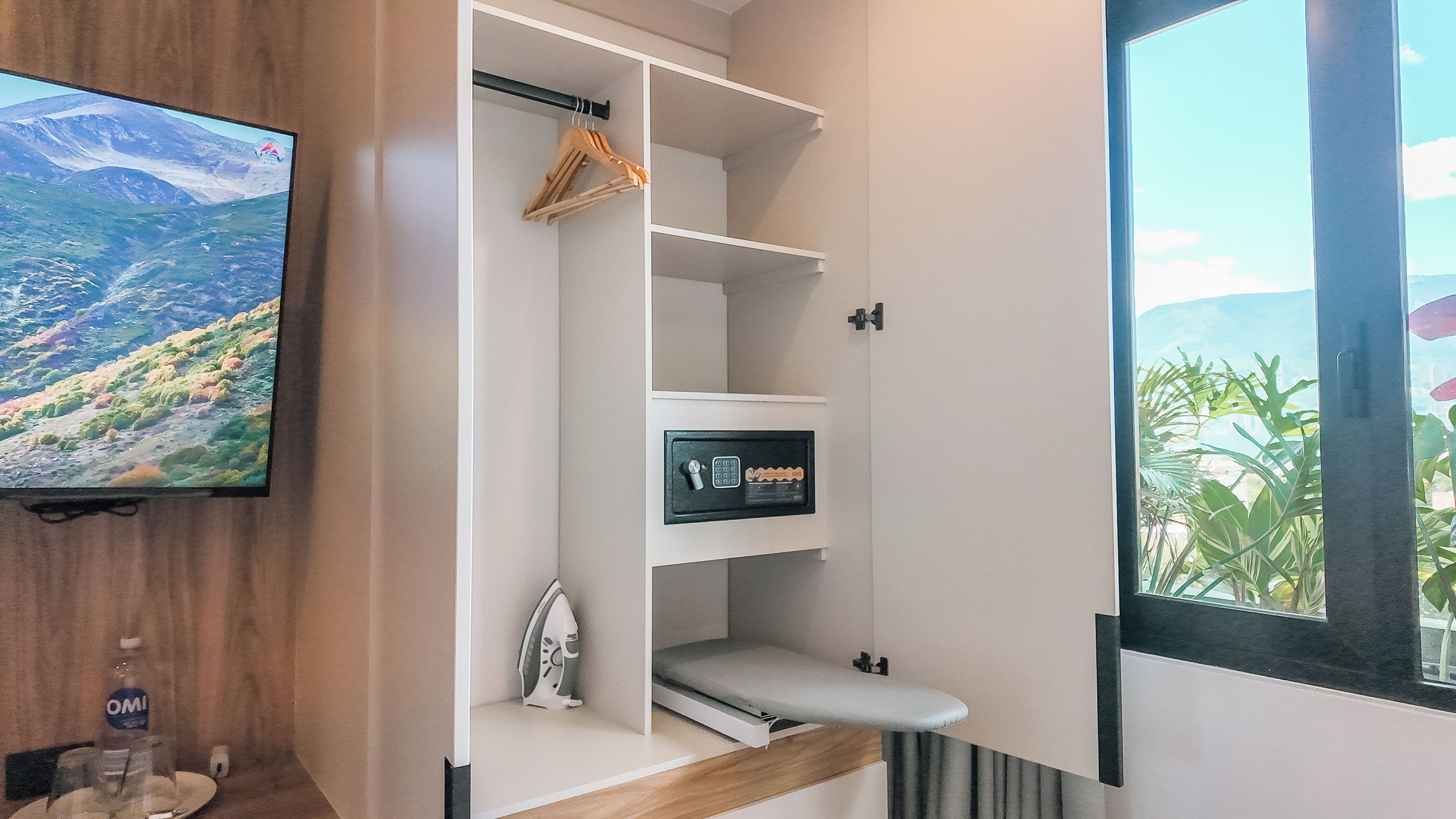 Ein Hotelzimmer mit integriertem Fernseher, einer Garderobe mit Kleiderhaken, einem kleinen Bügelbrett und einem Fenster mit Bergblick.
