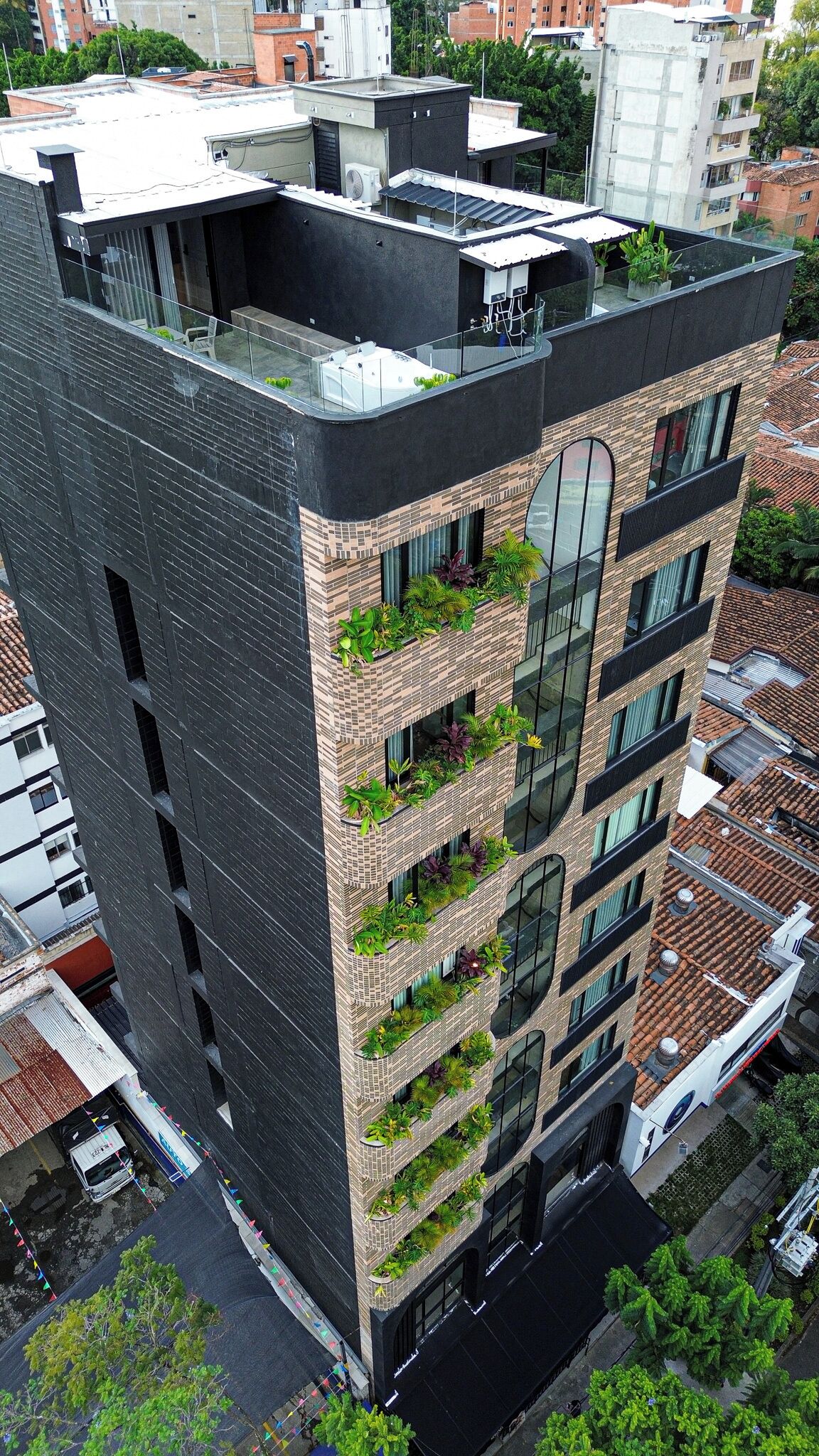 A modern multi-story building with a rooftop terrace, balconies with greenery, and a mix of brick and glass facade.