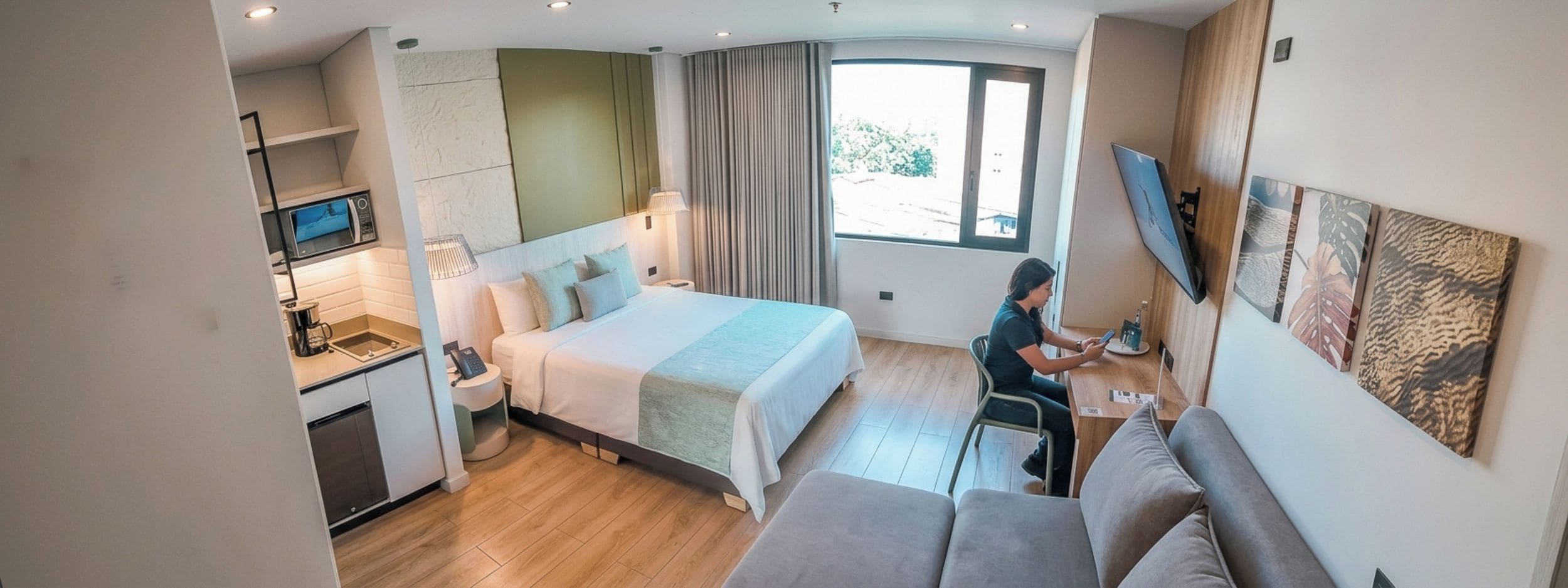 A compact hotel room with a bed, a small kitchenette, a seating area, and a TV mounted on the wall.