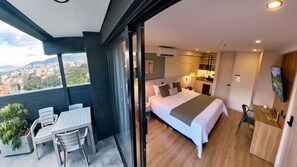 A modern bedroom with a large bed, a small kitchenette, and a balcony with a table and chairs.