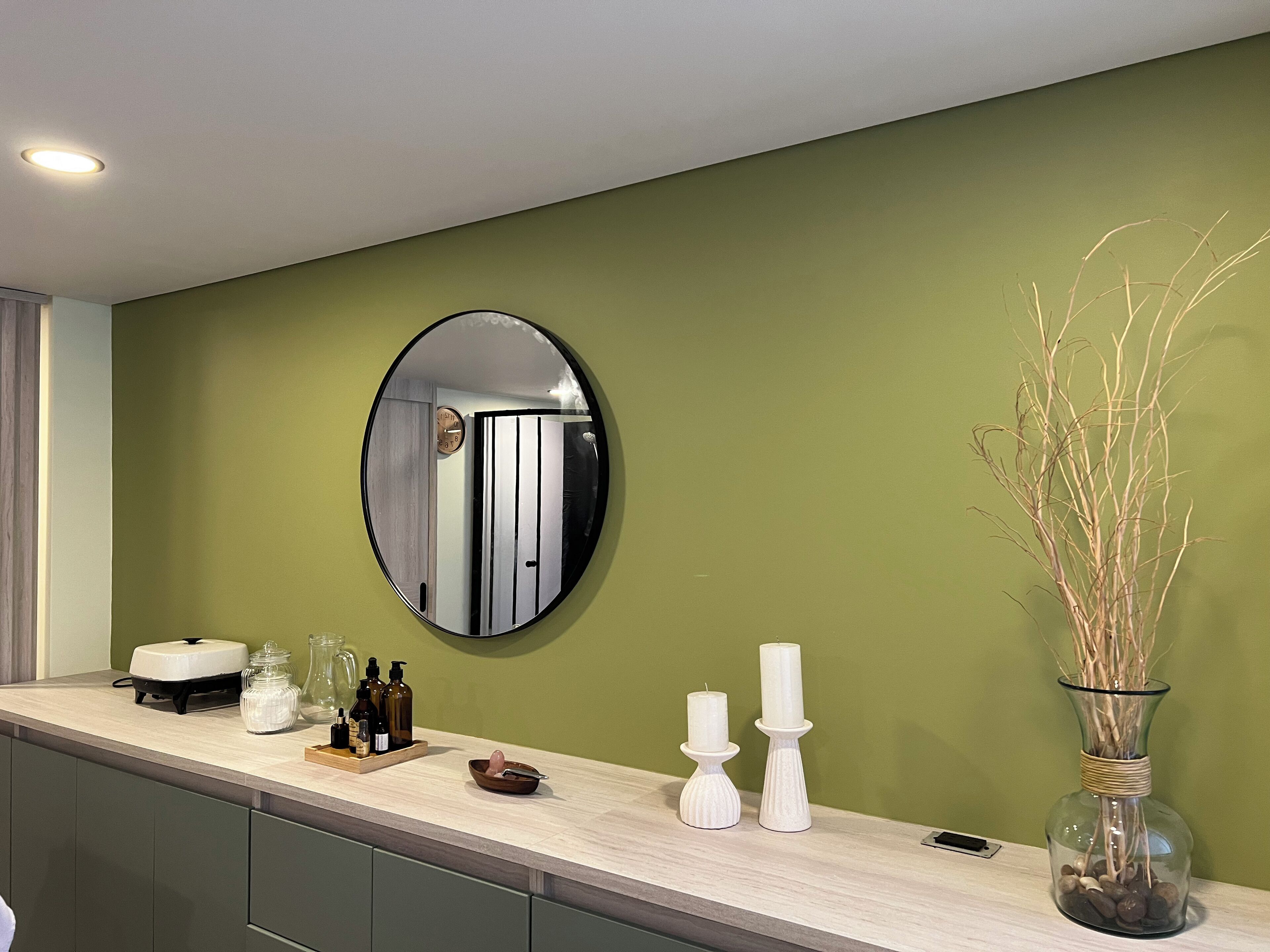 A modern bathroom with a green wall, a round mirror, a sink with toiletries, and a vase with reeds.