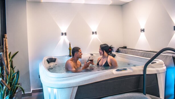 Un couple dans un jacuzzi, une bouteille de vin à portée de main, entouré d’un éclairage mural moderne et d’une plante en pot.