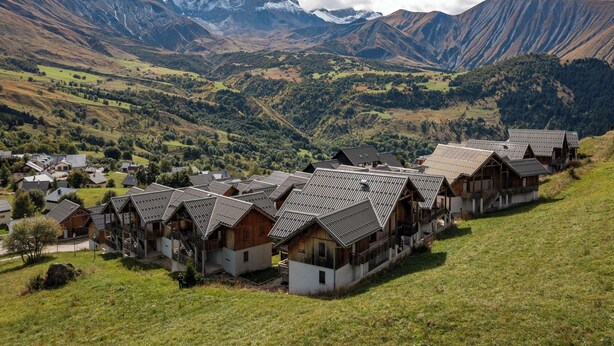 A cluster of traditional wooden houses with sloped roofs, set against a backdrop of mountains and a valley.