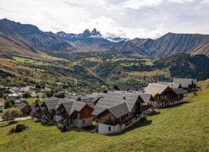A cluster of traditional wooden houses with sloped roofs, set against a backdrop of mountains and a valley.