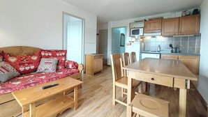 A compact living space with a sofa, wooden dining table, and chairs. A kitchen area with cabinets and a microwave is visible.