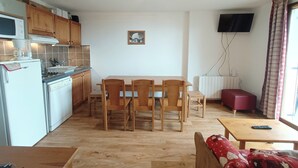 A compact kitchen with wooden cabinets, a dining table with chairs, a red sofa, and a TV mounted on the wall.
