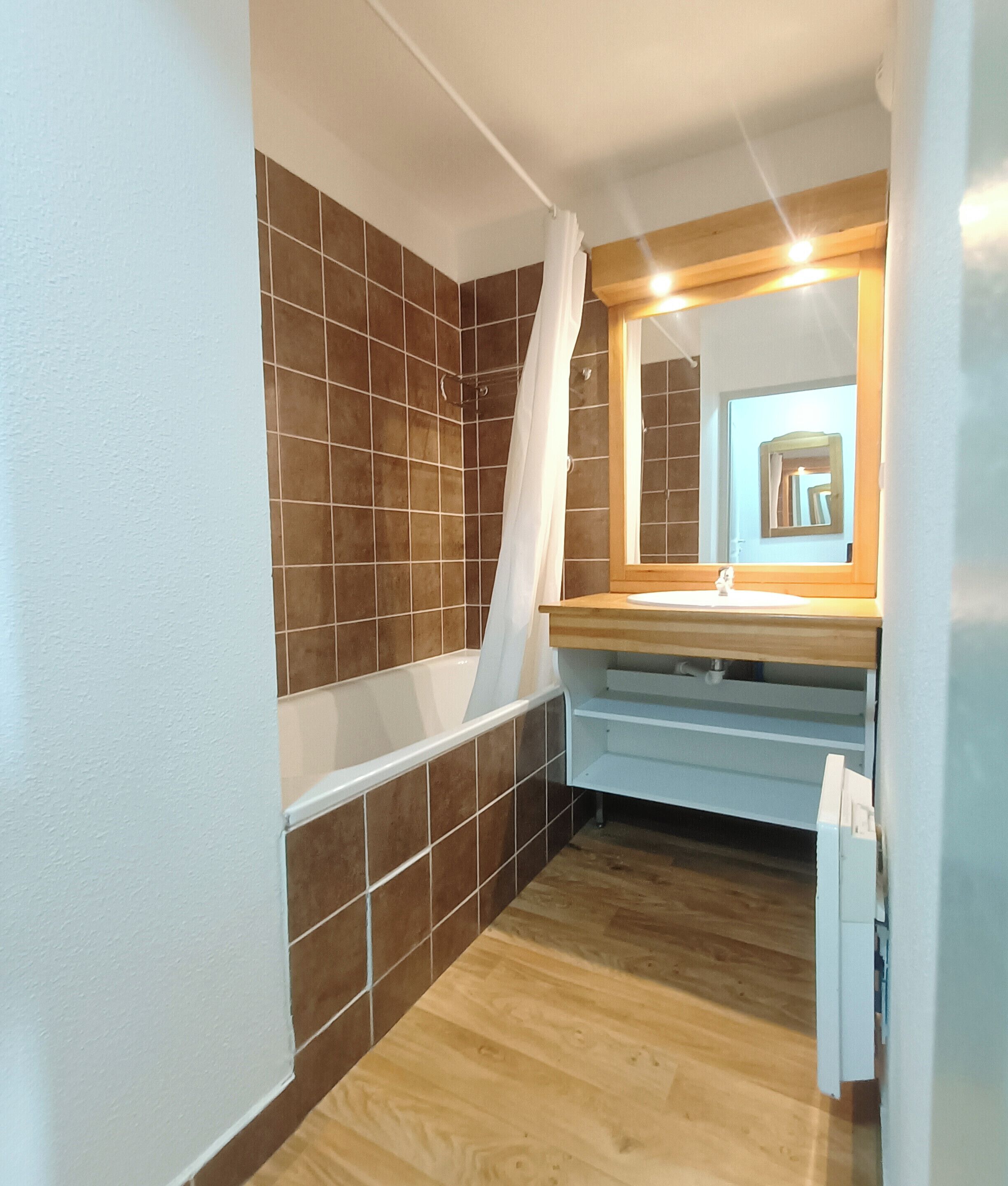 A bathroom with brown tiled walls, a wooden floor, a white bathtub, a wooden vanity with a mirror, and a white shower curtain.