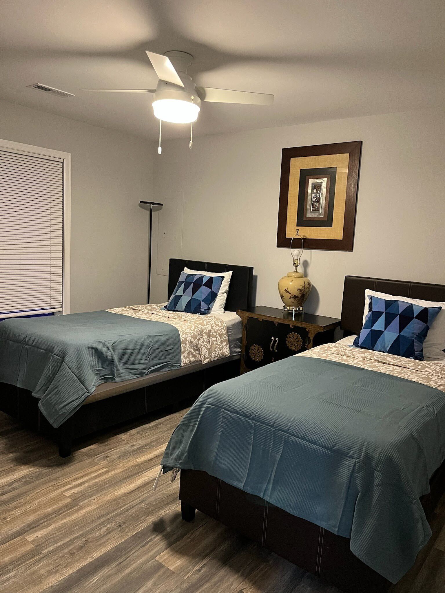 Una habitación con dos camas, cada una con cubrecama y almohadas a juego. Hay un ventilador de techo, una lámpara de pie y un cuadro en la pared.