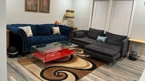 Een woonkamer met een blauwe bank, een rode salontafel en een grijze bank. Er hangen twee ingelijste foto's aan de muur, een plafondventilator en een raam met jaloezieën.