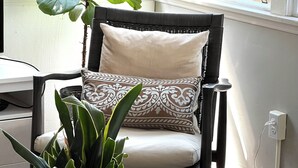 A room with a wicker chair, a potted plant, a tray with a remote control, and a book on a wooden table.