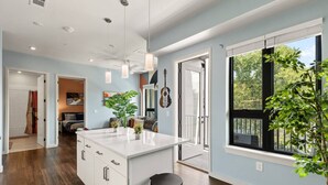 A modern kitchen with a central island, wooden flooring, and large windows offering views of the outdoors. - Pool, Rooftop Deck, City View, Gym + Gated Garage (Nashville)