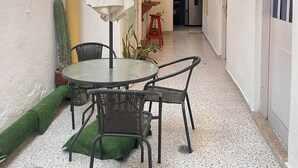 A narrow outdoor corridor with a glass ceiling, a round table with chairs, potted plants, and a red stool.