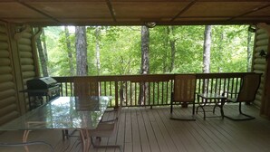 A covered outdoor deck with a glass-top table and chairs, a grill, and a view of a forested area.