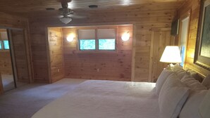 A bedroom with a large bed, wooden walls, two windows with shutters, a ceiling fan, and a nightstand with a lamp.