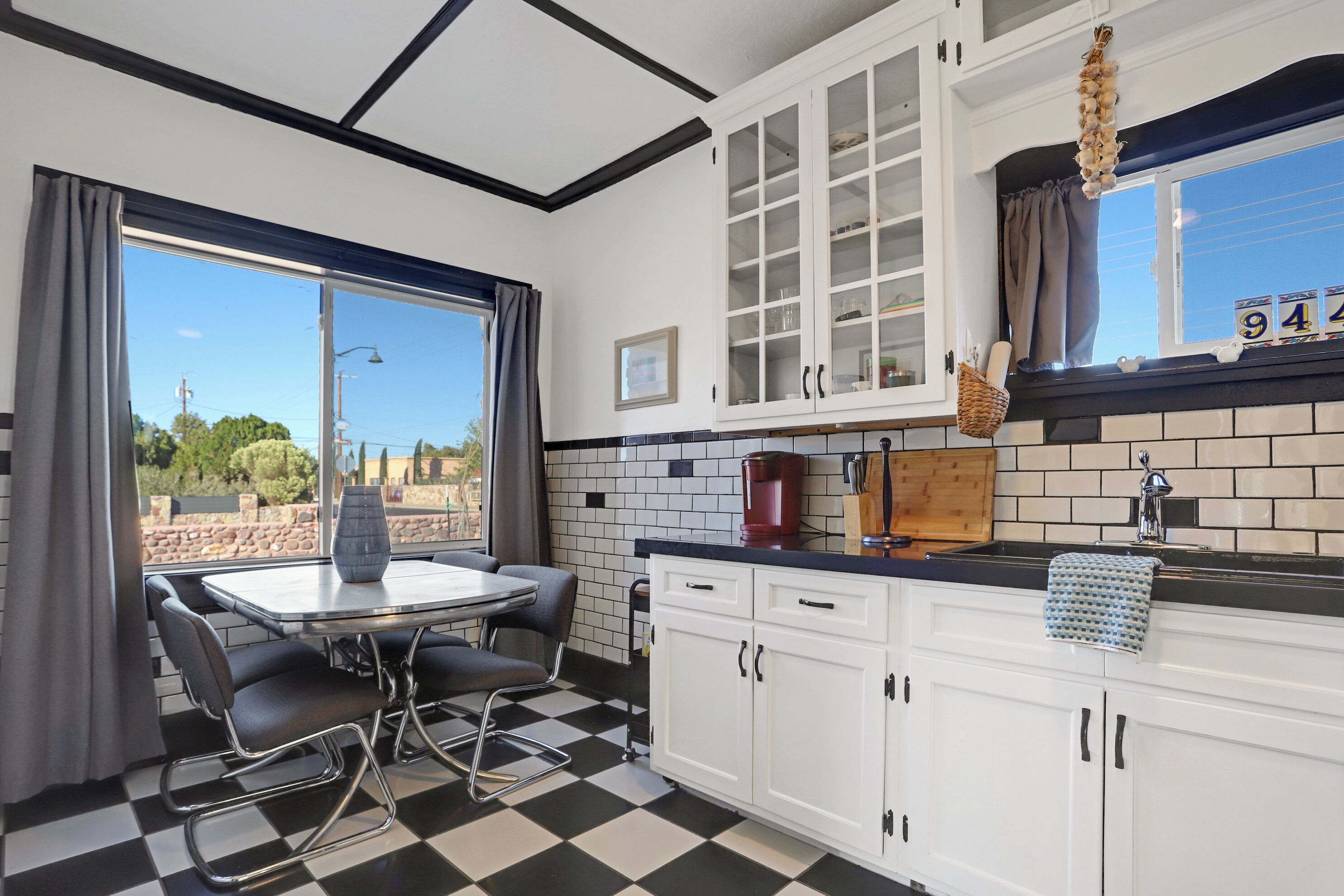 A kitchen with a checkered floor, white cabinets, a dining table with chairs, and a window with curtains.