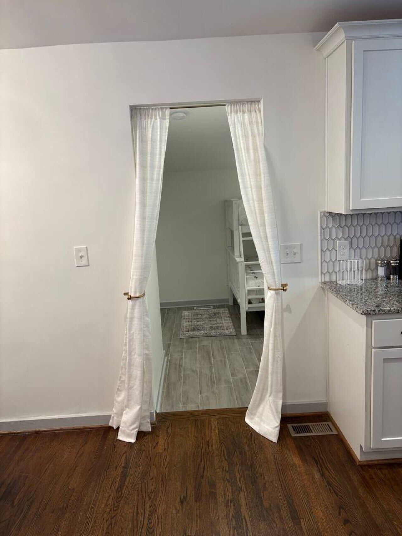 A room with a wooden floor, white walls, a kitchen area with a granite countertop, and a doorway with sheer curtains.